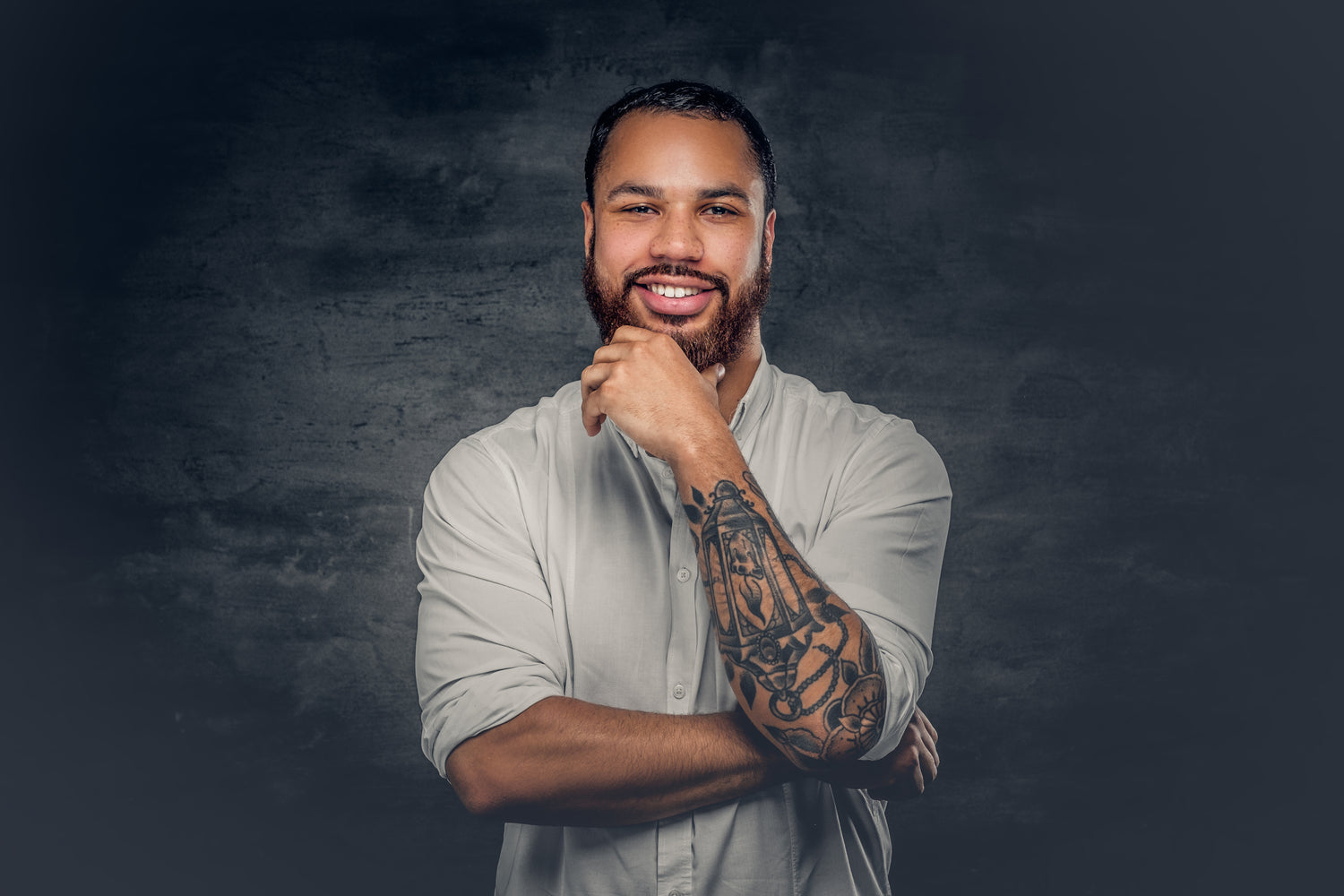 Man with tattoos on his arm wearing a light gray shirt against a dark background