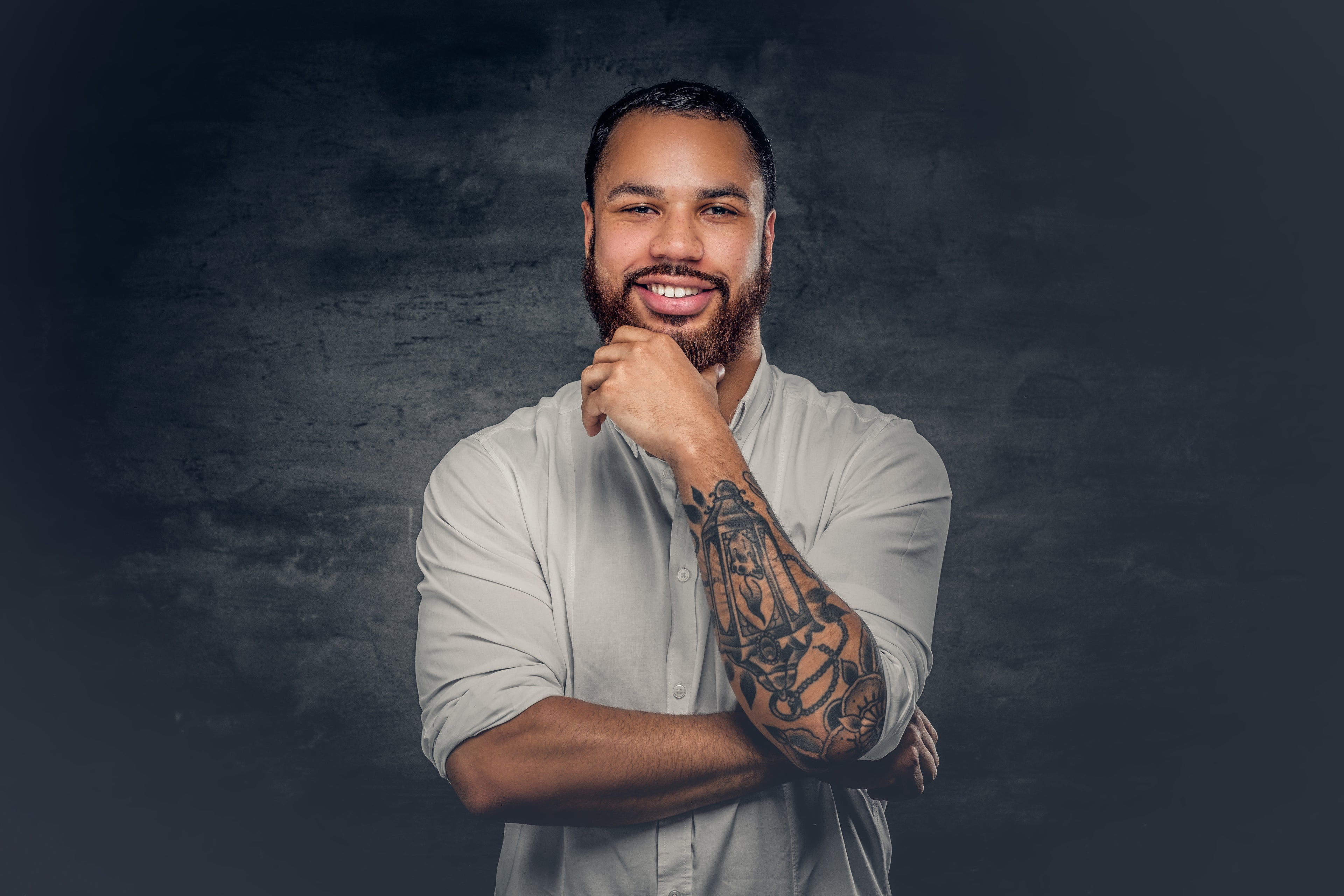 Man with tattoos on his arm wearing a light gray shirt against a dark background
