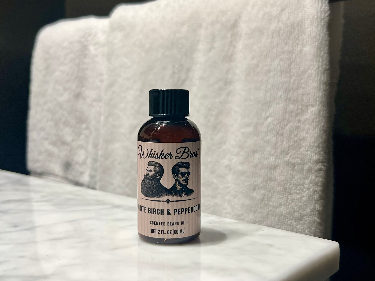 Bottle of Whisker Bros. White Birch & Peppercorn scented beard oil on a marble surface with a white towel in the background.