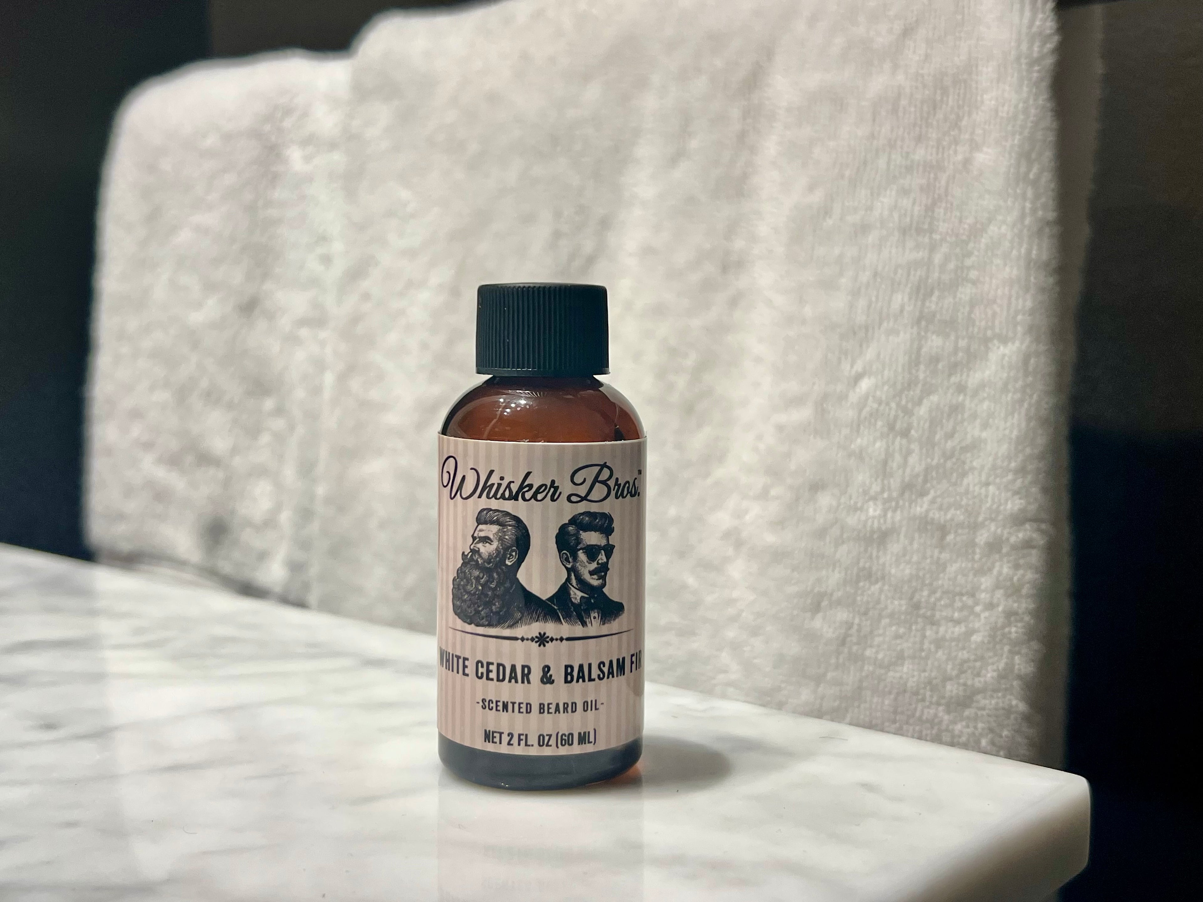 Bottle of Whisker Brand White Cedar & Balsam Beard Oil on a marble surface with a towel in the background.
