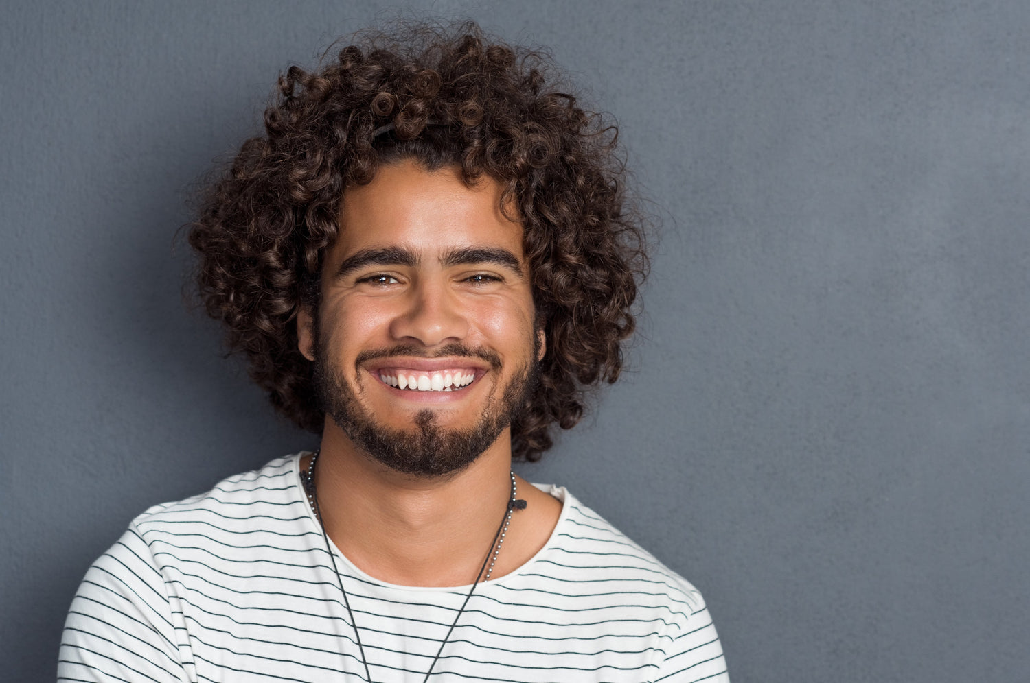 Man with curly hair and a beard wearing a striped shirt against a gray background