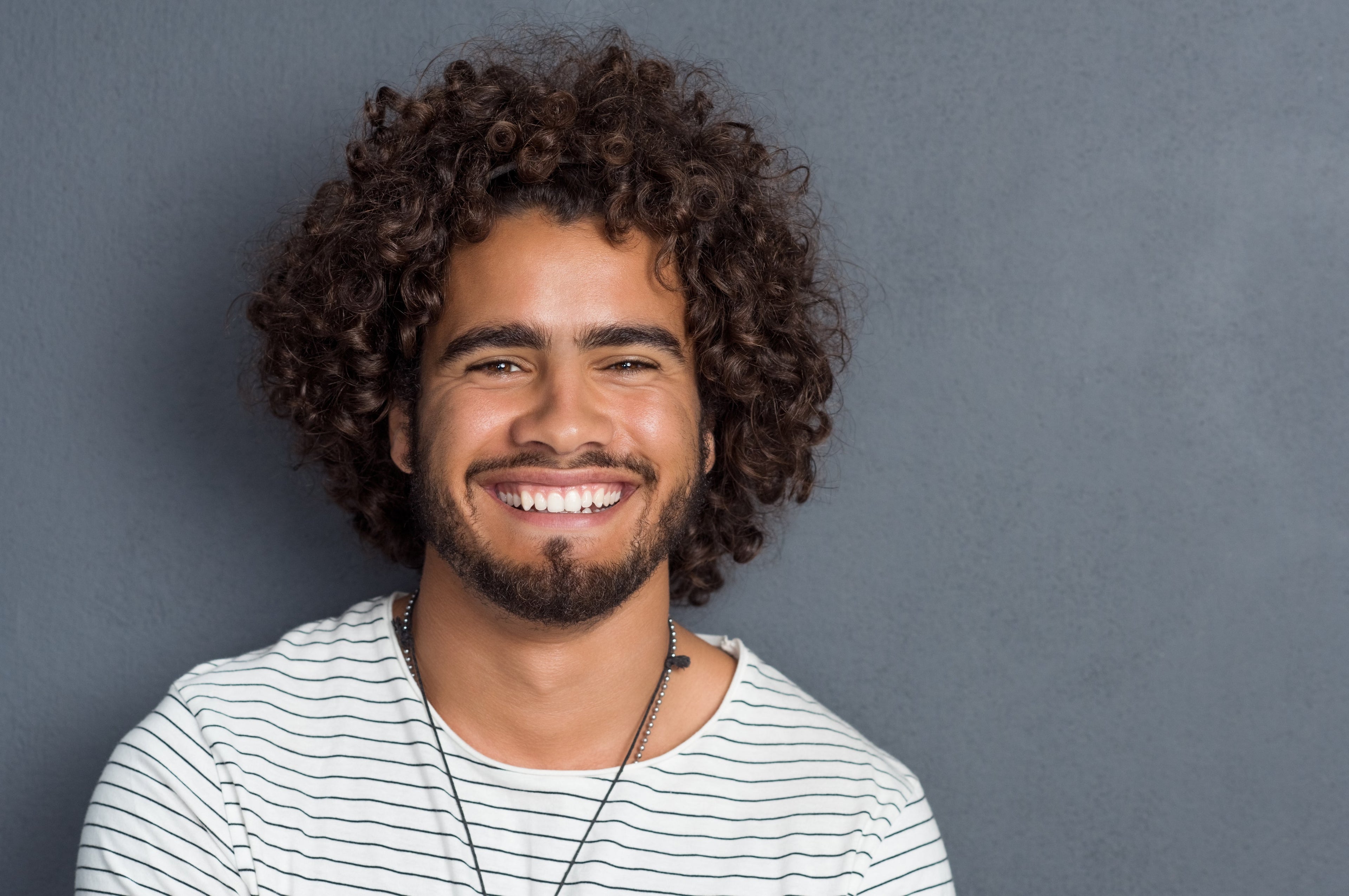 Man with curly hair and a beard wearing a striped shirt against a gray background