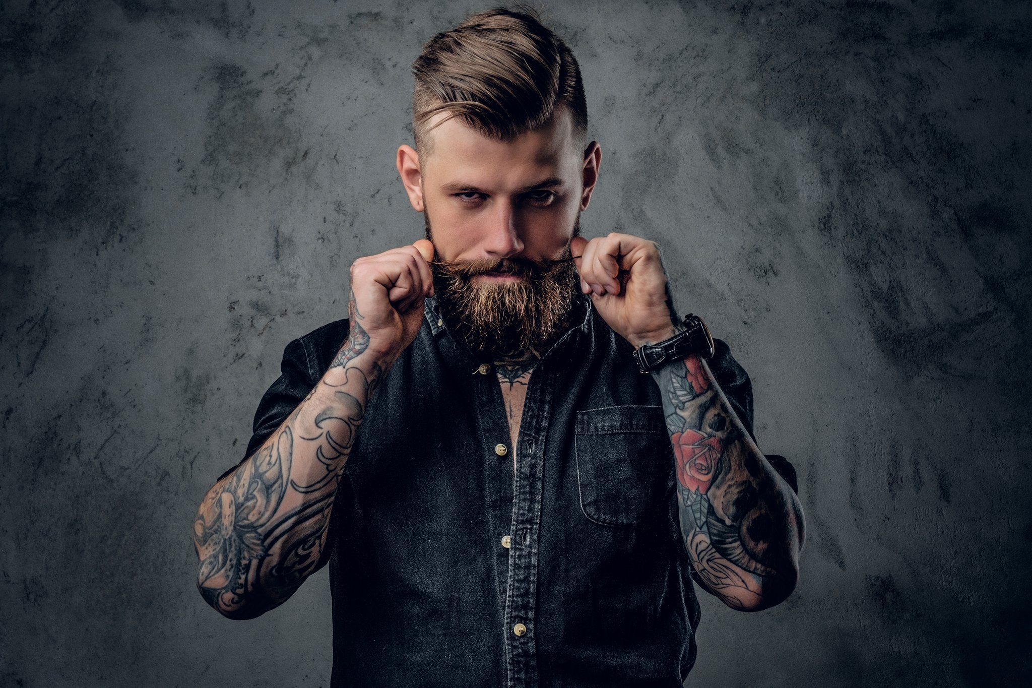 Man with tattoos and a beard holding a leather belt against a dark background